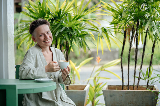 A woman with a short hairstyle and a smile is sitting in a cafe on the street drinking coffee with milk and enjoying her vacation.