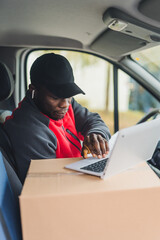 Young adult black delivery guy wearing work uniform sitting in driver seat of van working on laptop...