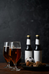 Two glasses with beer, bottles, snacks and soccer ball on wooden table.