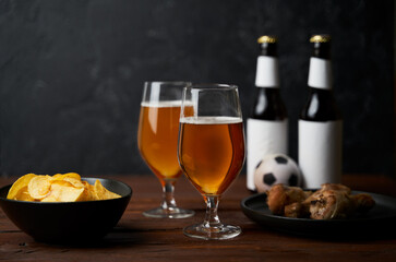Two glasses with beer, bottles, snacks and soccer ball on wooden table.