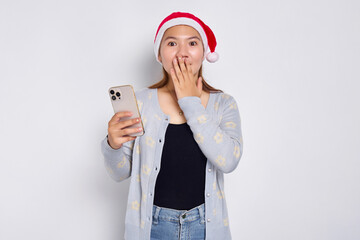 Surprised expression Young attractive Asian woman in a Christmas hat holding mobile phone and covering her mouth with hand isolated on white background