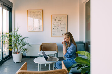 Side view of a woman working at the bright office, looking through the window, drinking coffee.