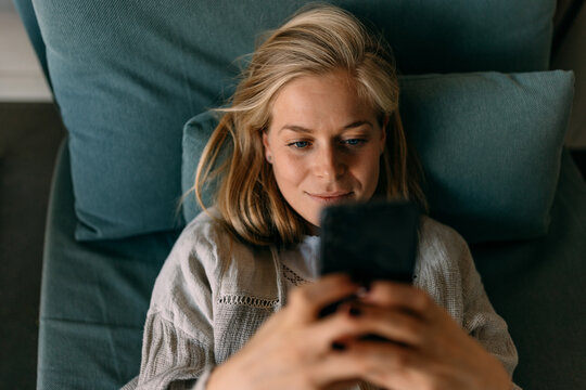 Blonde female enjoying herself alone, laying on the couch and using a phone.