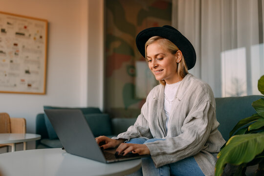 Smiling Woman Typing Something Over The Laptop, Dressed Casually, Wearing A Hat.