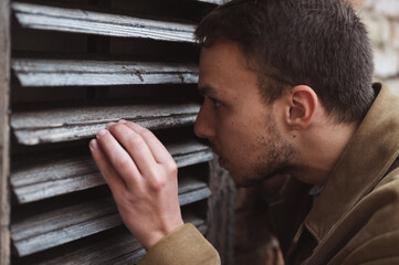 Obraz premium Suspicious looking young man peeking from outside trough wooden window blinds. Stalker, close up.