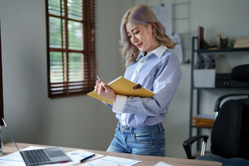 Portrait of a young Asian woman showing a smiling face as she using notebook, computer and financial documents on her desk in the early morning hours
