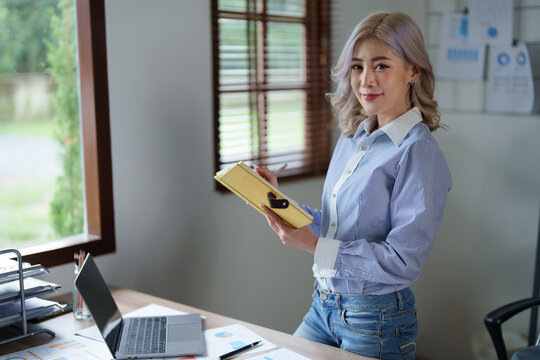 Portrait of a young Asian woman showing a smiling face as she using notebook, computer and financial documents on her desk in the early morning hours