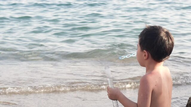 boy kid doing soap bubbles against sea background water waves horizont line.adorable naket child caucasian baby playing on sands beach vacation family together many balloons flying slow motion