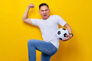 Excited young Asian man 20s wearing a white t-shirt supporting a football sports team, holding in...