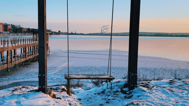 An empty wooden swing moving outdoors, without people. Beautiful view of the pond covered with snow and ice against the background of the smoking pipes of the Topol-M rocket factory. Russia
