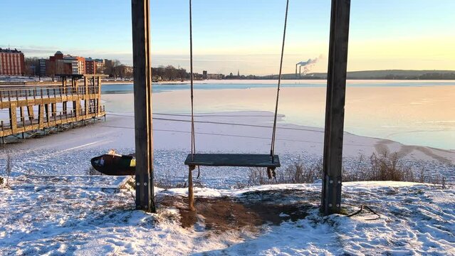 An empty wooden swing moving outdoors, without people. Beautiful view of the pond covered with snow and ice against the background of the smoking pipes of the Topol-M rocket factory. Russia
