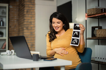 Beautiful young pregnant woman looking at ultrasound pictures of her baby. Businesswoman having video call