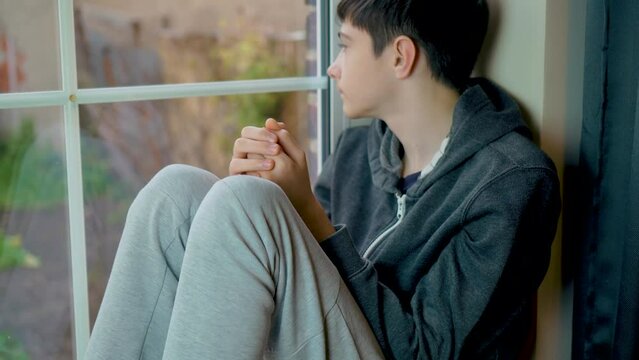 Sad boy refugee from Ukraine sitting looking through window and cannot go outside home because Russian military invasion. Save lifes, no war, world peace, stop conflict, born love. Childhood in