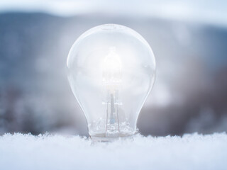 Close-up of a lit light bulb sticking up from soft snow with a blurred winter landscape in the background