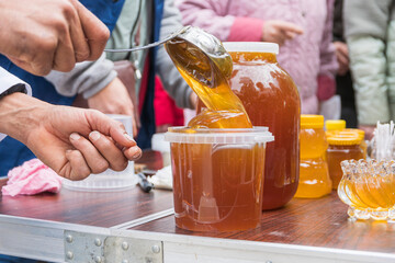 Autumn agricultural fair. Male hands, using ladle, put thick orange-brown mass of bee honey into plastic container. Sale of bee products.