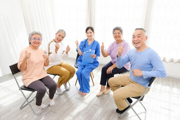 The caregiver therapist sits with a group of Asian senior people with thumbs up in a circle for...