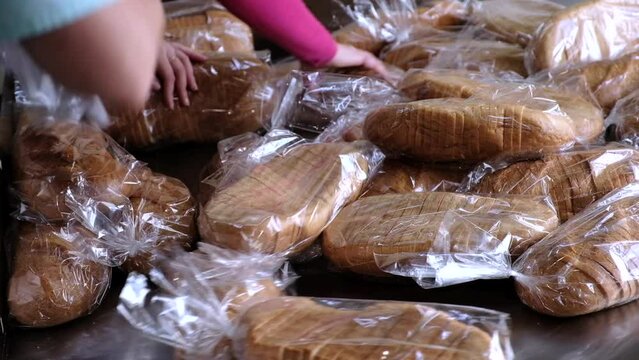 Process of sorting freshly baked white bread on conveyor in bakery. Female hands of workers put sliced loaves of white bread in transparent plastic bags closeup