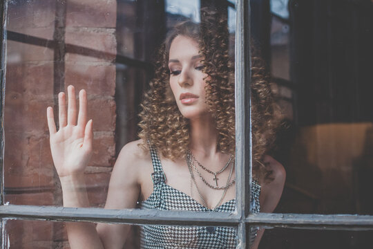 Portrait of beautiful pensive woman with curly hait and touching window glass