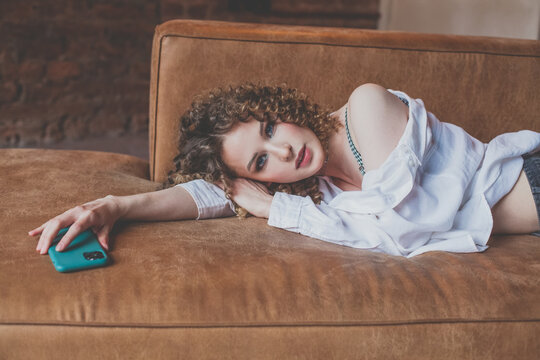 Beautiful glamorous woman in white shirt with curly hair lies on the sofa with green mobile phone in her hand