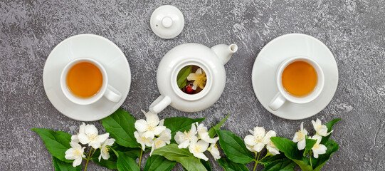 Glass white cups and teapot of hot green tea with spring jasmine flowers and blooming branches on dark blue texture background. Phytotea, healthy food