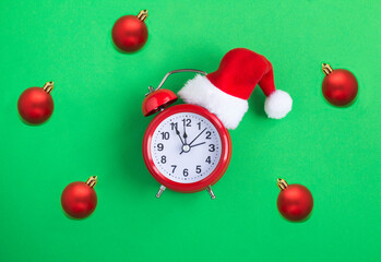 Red alarm clock with Christmas Santa hat and red bead on the green background. Close-up. Copy space.