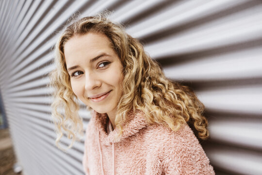 Young blonde girl stands in front of metal wall and looks to the side smiling