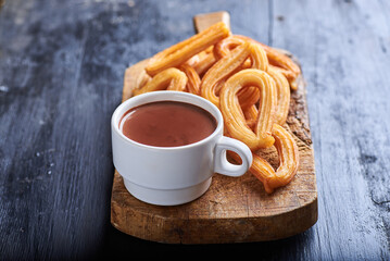 Chocolate en una taza de color blanco sobre una superficie rústica y rodeada de churros y un fondo negro