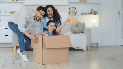 Hispanic Indian multiracial multiethnic family homeowners buyers of real estate loving happy parents pushing cardboard box with daughter child girl sitting inside having fun play ride game moving day