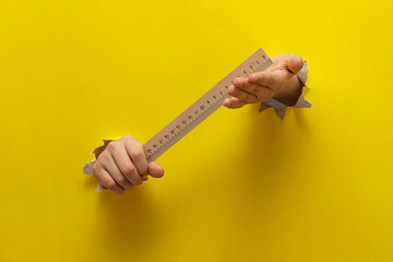 Two children's hands stretched through a torn sheet of yellow paper hold the student's ruler
