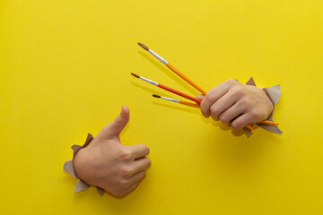 Two children's hands stretched through a torn sheet of yellow paper hold brushes for drawing