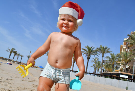 Portrait of a cute toddler boy wearing Santa hat playing on the beach. Christmas vacation with kids concept