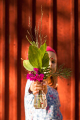 Child presents bouquet of flowers in front of red wooden wall. Girl proudly holds self-picked...