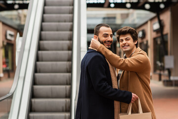 happy gay man in beige coat hugging neck of bearded boyfriend holding shopping bags near escalator.