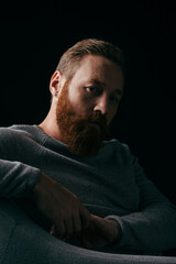 Stylish bearded man in jumper sitting on armchair isolated on black with shadow.