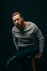 Trendy bearded man in grey sweater sitting on chair and looking at camera isolated on black.