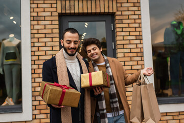 stylish gay couple holding Christmas gift boxes and shopping bags near shop with showcases on...