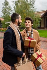 cheerful gay man holding Christmas presents near trendy boyfriend with shopping bags on blurred...