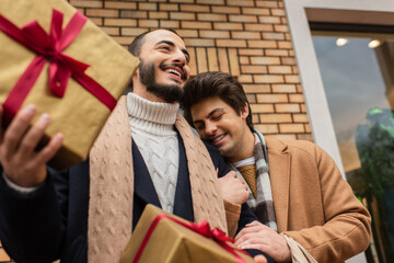 low angle view of bearded gay man holding gift boxes near happy boyfriend with closed eyes on street.