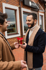 young gay man presenting small gift box to smiling bearded boyfriend near blurred shop on street.