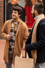 happy gay man in beige coat looking away near bearded boyfriend with shopping bags.