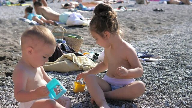Little siblings, brother and sister, three and one year old kids playing with pebbles on Black sea beach. Kid plays at sea on summer family vacation. Children sitting on the beach under the sun rays.