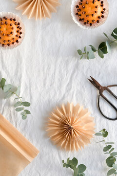 Fragrant pomander balls handmade from tangerines with cloves. Flat lay on off white textile tablecloth with aromatic wintertime green eucalyptus twigs. Old scissors and brown paper stars, snowflakes.