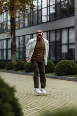 full length of bearded man in beanie hat and trendy sunglasses jumping while smiling near modern building.