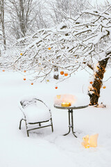 Snow covering apple tree in home garden in winter, decorated with lot of copper metallic Christmas baubles and warm white string led lights illuminated. Table with chair, wine glass and outdoor candle
