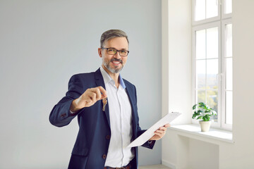 Portrait of friendly male real estate agent showing key to house that you can buy or rent. Smiling Caucasian middle-aged man in business suit posing with clipboard and key in bright room.