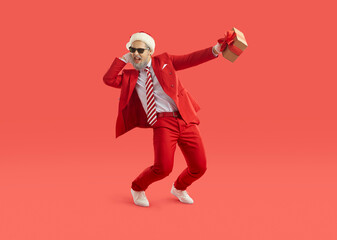 Cool energetic man in santa hat having fun and dancing with christmas gift in hand. Man with gray beard wearing red suit and sunglasses is ready for Christmas party. Isolated on red background. Banner