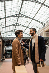 cheerful gay men in trendy outfits holding shopping bags and looking at each other under transparent roof on street.