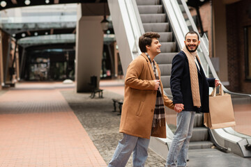 cheerful gay couple in fashionable outfit holding hands while standing with shopping bags near...