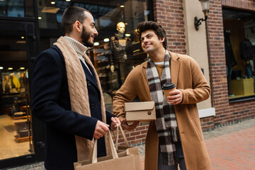 happy gay man with shoebox and takeaway drink looking at bearded boyfriend with shopping bags near blurred store.