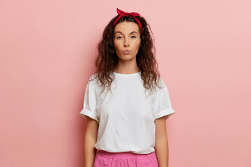 Young curly haired woman, dressed in white t-shirt, and red bandana on the head, keeps lips folded sends air kiss, isolated over pink wall